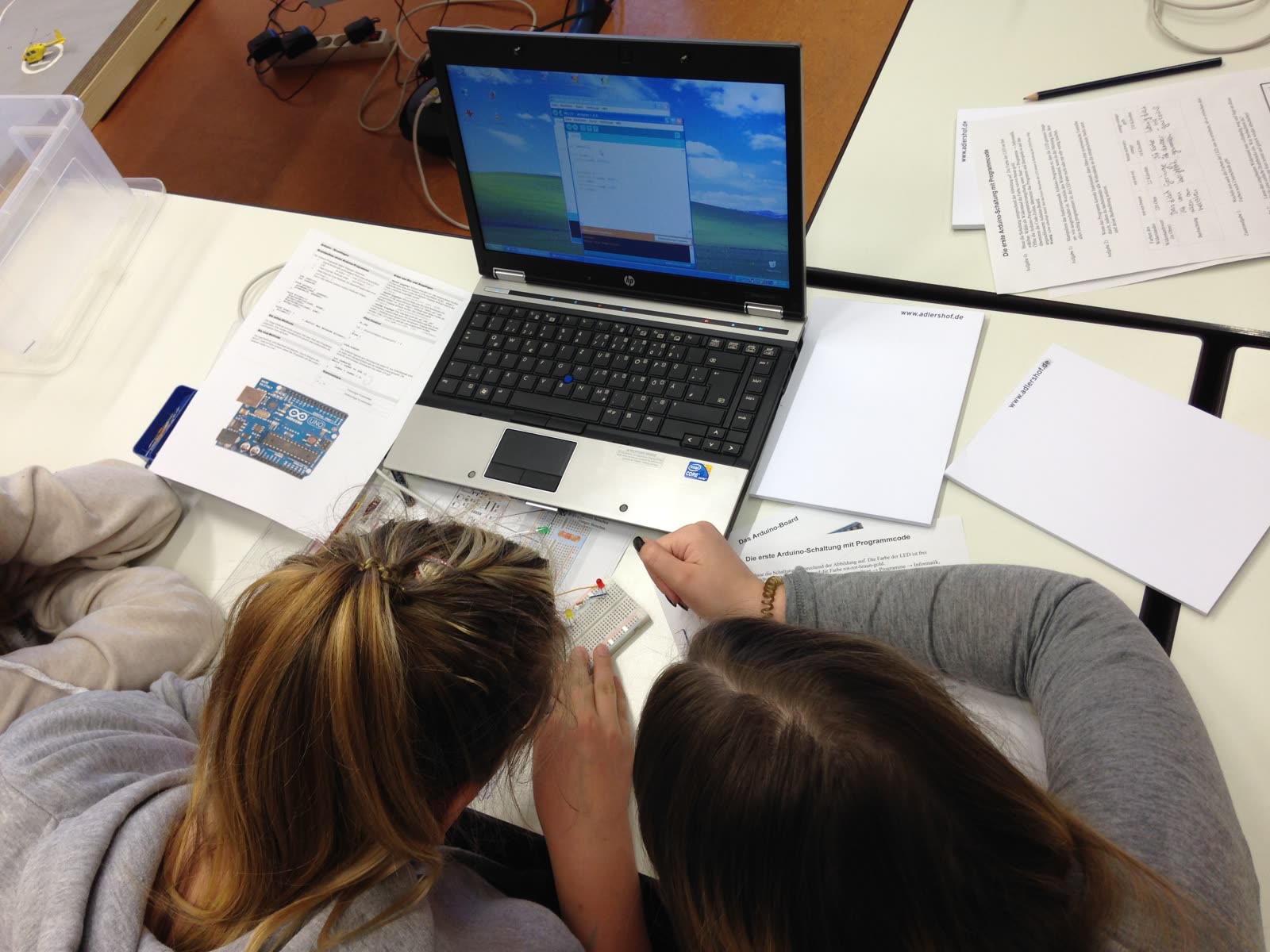 Zwei Schüler:innen von hinten am Laptop, daneben Arbeitsblätter und Bauteile