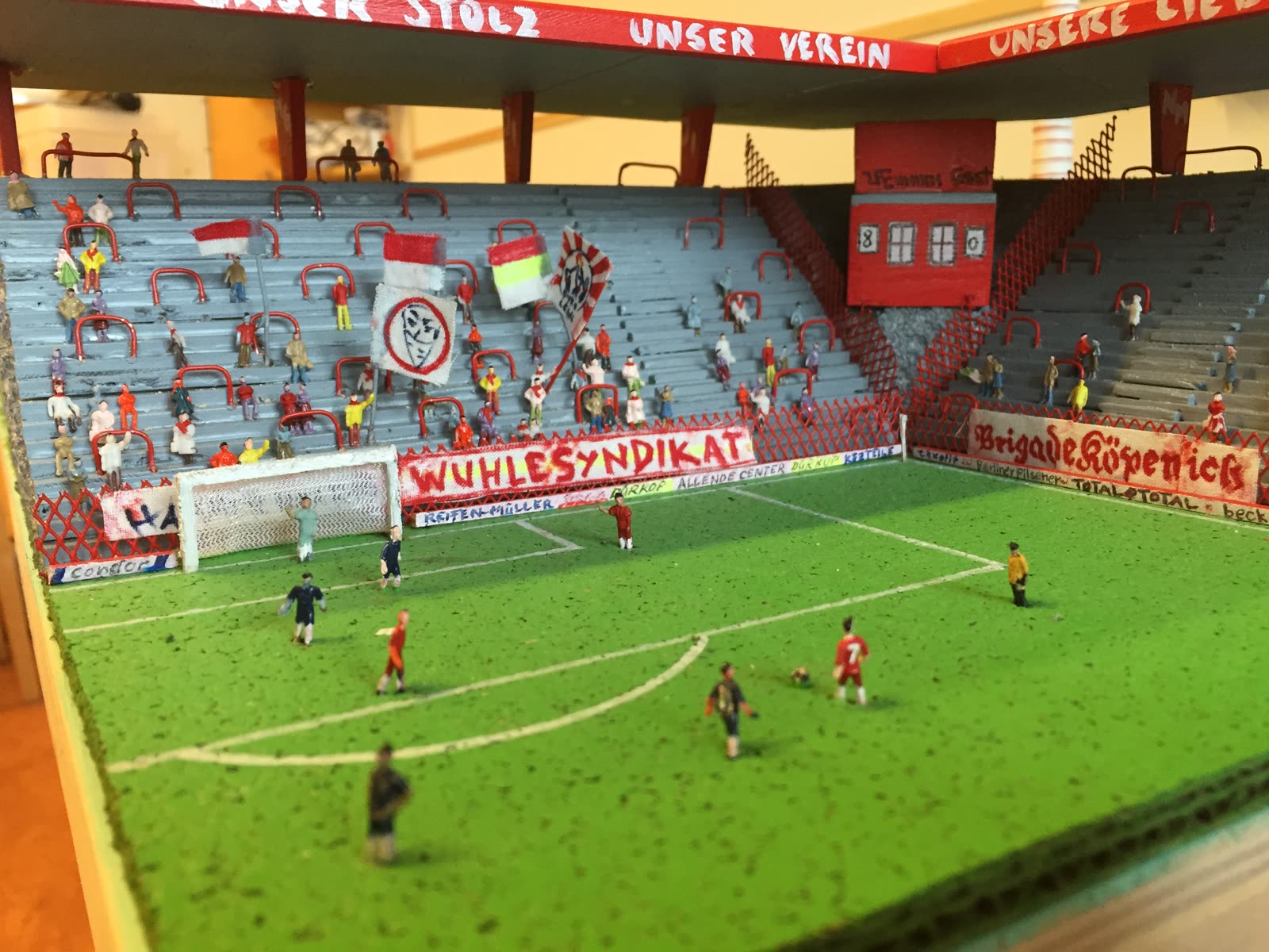 Modell-Fußballstadion mit Spielern auf dem Rasen und voller Fankurve mit Fahnen und Bannern
