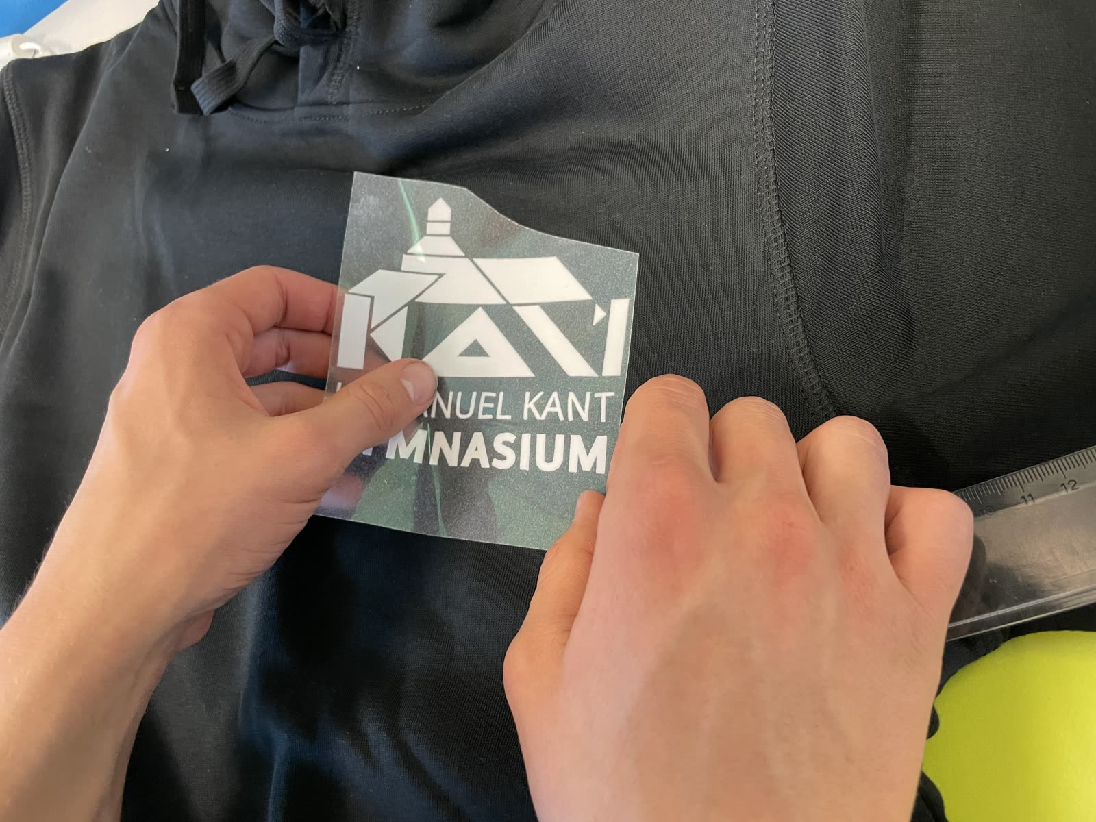 Hände richten ein weißes Kant-Logo aus Flexfolie auf einem dunklen Hoodie aus, bevor es aufgebügelt wird
